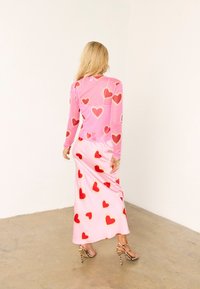 Γυναίκα με μακριά ξανθά μαλλιά που φοράει ροζ φόρεμα και τοπ με κόκκινα μοτίβα καρδιών, στέκεται σε τσιμεντένιο δάπεδο μπροστά σε λευκό τοίχο.