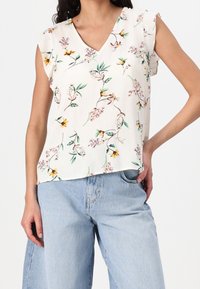 Nainen, joka on pukeutunut valkoiseen hihattomaan puseroon, jossa on keltainen ja vaaleanpunainen kukkakuosi, sekä vaaleansiniset korkea vyötäröiset leveät housut, käsi taskussa.