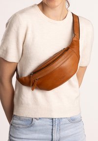 Vrouw met een beige leren crossbodytas over een crèmekleurige korte mouwen trui en lichtblauwe jeans tegen een neutrale achtergrond.