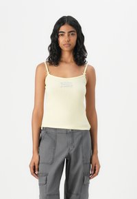 Yellow ribbed tank top with thin straps, featuring a subtle blue graphic at the center. Paired with gray cargo pants.