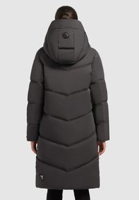 Sieviete, kas ģērbusies garā melnā ziemas jakā ar lielu kapuci, redzama no mugurpuses pret vienkāršu gaišu fonu.