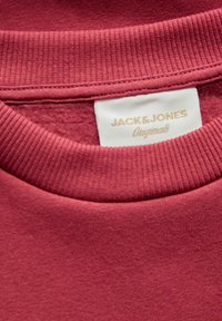 Bordeauxrode trui met geribbelde kraag. Label met de tekst "JACK & JONES Originals" in goud op een gestructureerde ivoorkleurige achtergrond.