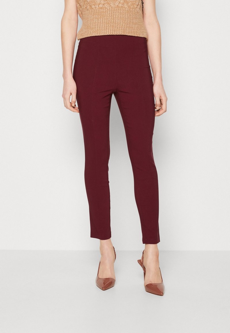Femme portant un pantalon bordeaux ajusté, un haut beige en maille sans manches et des escarpins marron à bout pointu, debout devant un fond blanc uni.