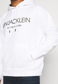 Valkoinen huppari, jossa on etutasku ja nyörikiinnitys. Edessä mustalla tekstillä "VIVIACKLEIN EST 1968, NEW YORK".