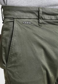 Olivgrüne Freizeithorts aus einem glatten Stoff, ausgestattet mit einem silbernen "GUESS"-Logo am Bund und seitlichen Taschen.