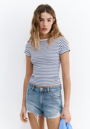 Jeune femme aux cheveux ondulés portant un t-shirt ajusté rayé blanc et bleu marine et un short en jean usé tenant un tissu bleu.