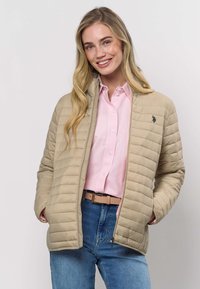 Bēšs dūnu žakete ar rāvējslēdzēja priekšpusi un augstu apkakli, valkāta virs gaiši rozā krekla. Kreisajā krūšu pusē ir neliels logotips.