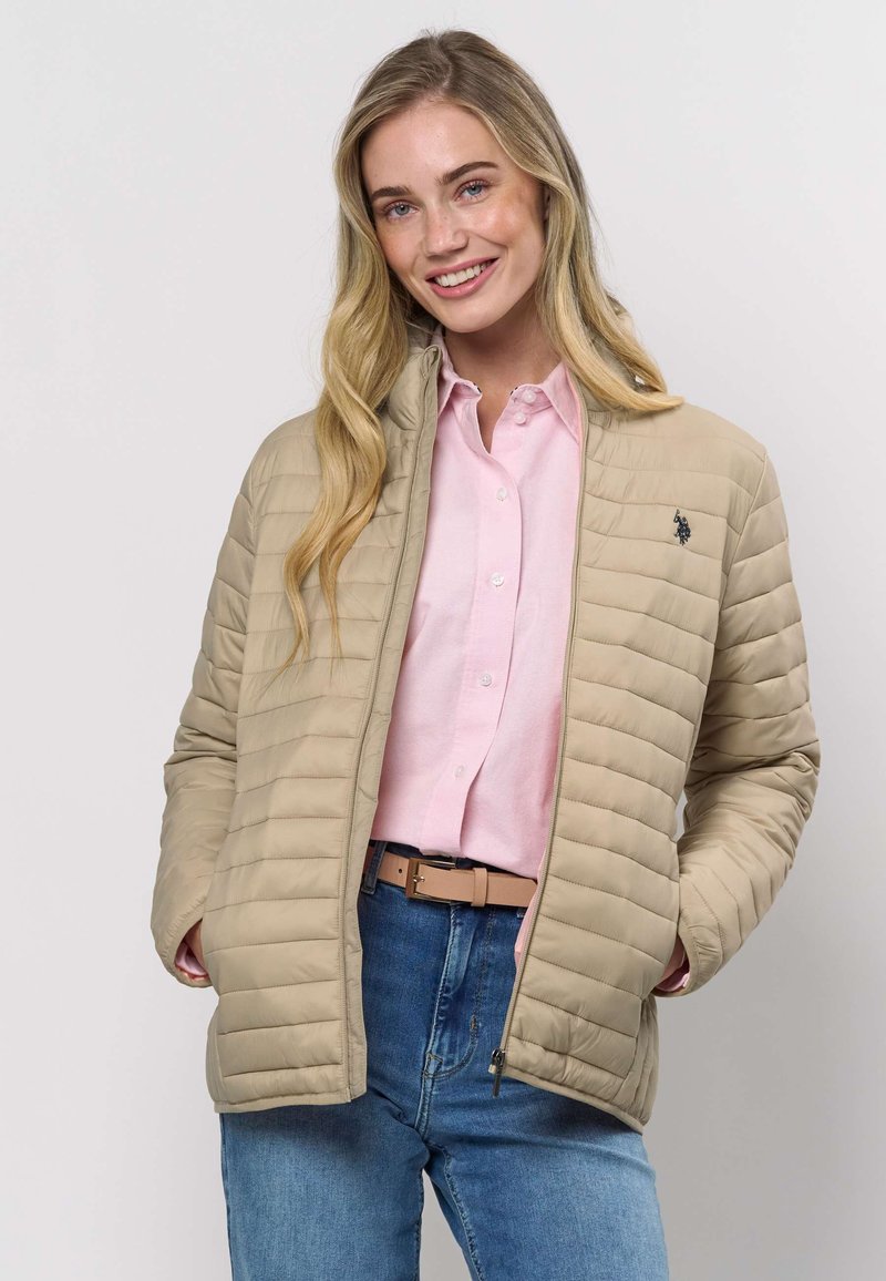 Bēšs dūnu žakete ar rāvējslēdzēja priekšpusi un augstu apkakli, valkāta virs gaiši rozā krekla. Kreisajā krūšu pusē ir neliels logotips.