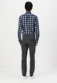 Mann som står med ryggen til iført en marineblå og hvit rutete skjorte, mørkegrå jeans og hvite joggesko mot en hvit bakgrunn.