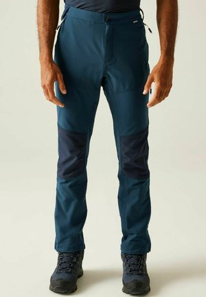 Man wearing blue outdoor pants with reinforced dark knee patches and hiking boots, standing against a plain background.