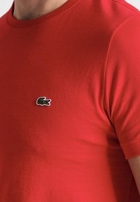 Camiseta de algodón roja con mangas cortas y cuello redondo, que presenta un pequeño logo bordado de cocodrilo verde en el lado izquierdo del pecho.