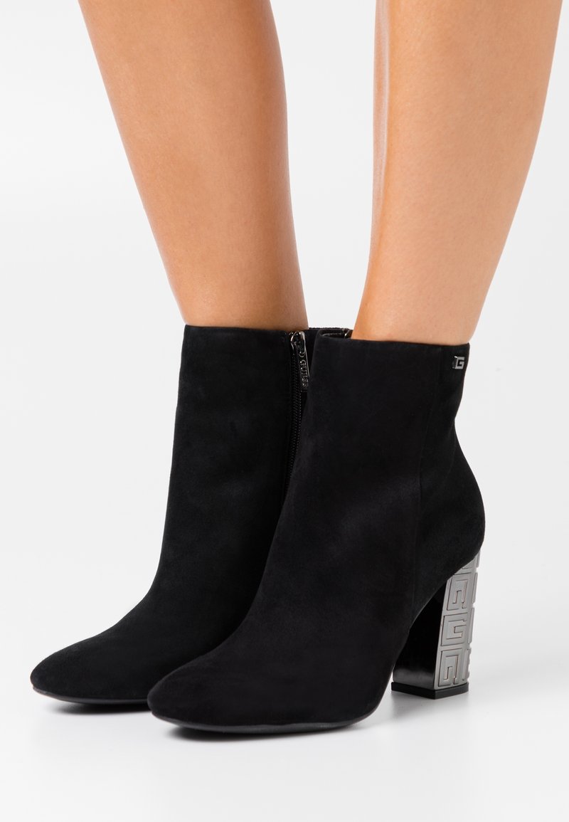 Botas de tobillo de ante negro con punta afilada y un tacón bloque metálico decorativo que presenta un patrón geométrico. Cierre con cremallera lateral.