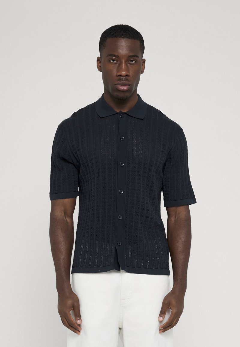 Polo noir à manches courtes avec boutons et rayures verticales texturées, porté par un homme debout en pantalon blanc sur un fond uni.
