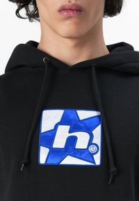 Black hoodie featuring a prominent blue and white embroidered logo on a square patch with a star design. Adjustable drawstrings included.
