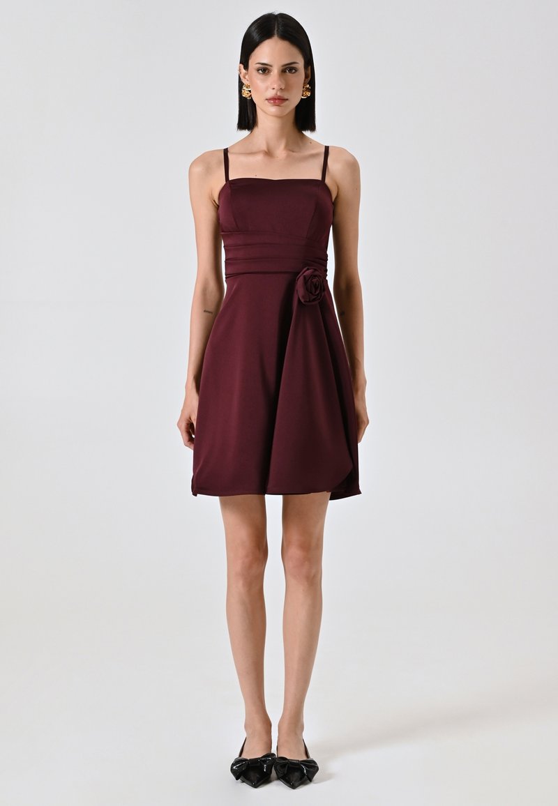 Bordeaux mouwloos jurkje met een fitted lijfje en zijdetails van bloemen. Zachte stof, A-lijn vorm, valt boven de knie. Zwarte schoenen met strikken.