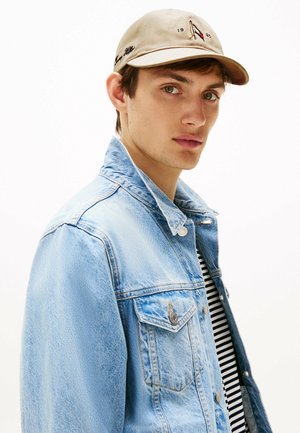 Light blue denim jacket with two front pockets, striped t-shirt underneath, and a beige cap with black embroidered details.