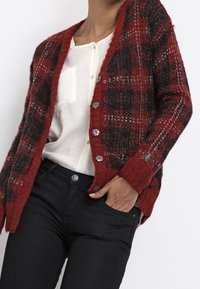 Cardigan de patrón rojo y negro con un tejido texturizado, que presenta un escote en V, cinco botones frontales y mangas largas con detalle en los puños.