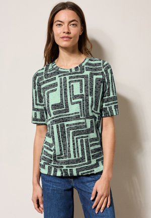 T-shirt vert à manches courtes avec un motif géométrique noir, fabriqué en tissu doux. Présente un col rond et une coupe décontractée.