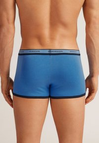 Herrblå kalsonger i trunk-modell gjorda av mjukt material, med ett kontrasterande svart midjeband med "intimissimi"-logotyp. Slät textur och enkel design.