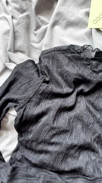 Black, long-sleeved top with a textured, rippled design. Features a scalloped neckline and soft, lightweight fabric against a gray background.