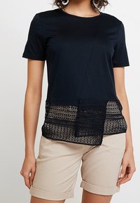 Modelka oblečená v černém tričku s texturovanou krajkou na spodním okraji, spárovaném s béžovými zkrácenými šortkami, na jednobarevném pozadí.