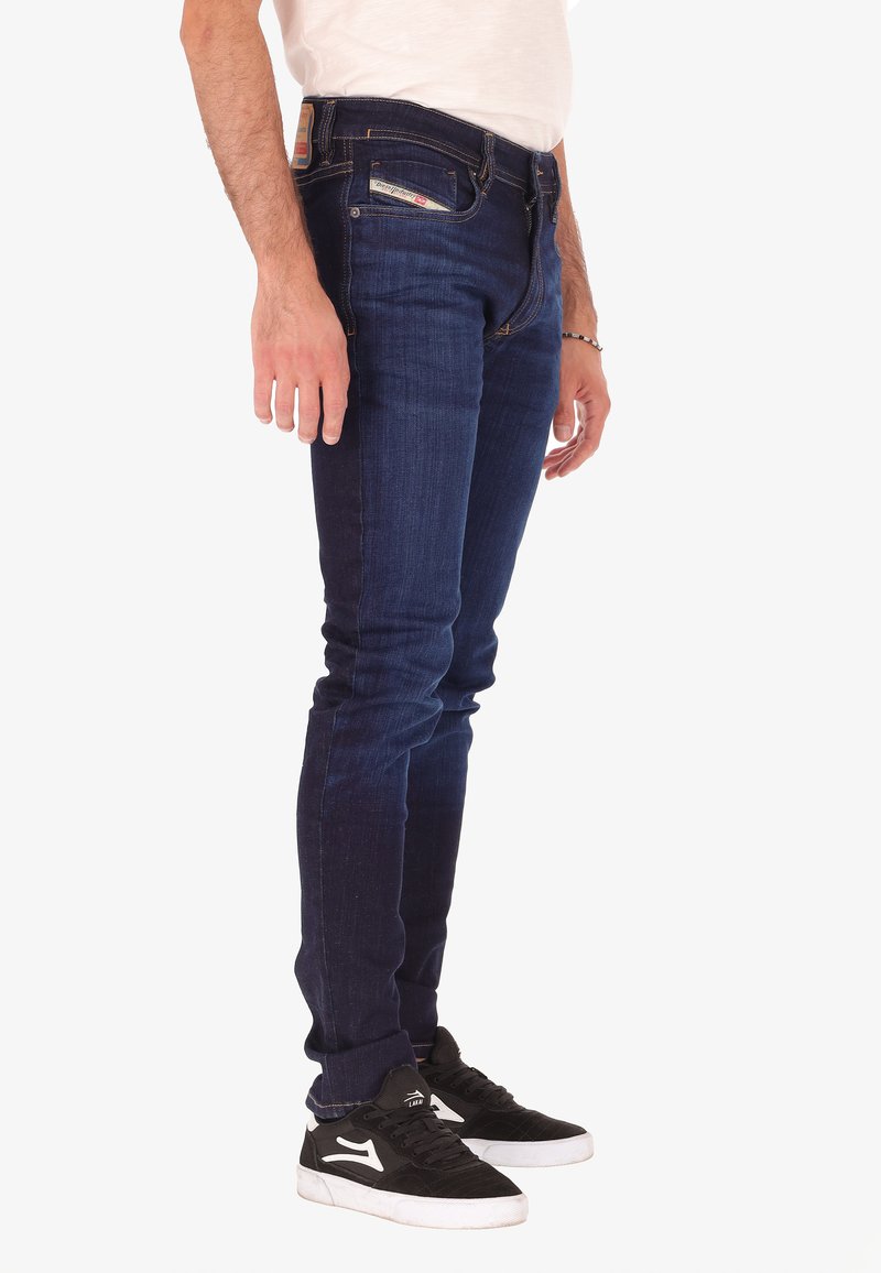 Homme portant un jean slim-fit bleu foncé et des baskets noires avec des semelles blanches, se tenant contre un fond blanc.