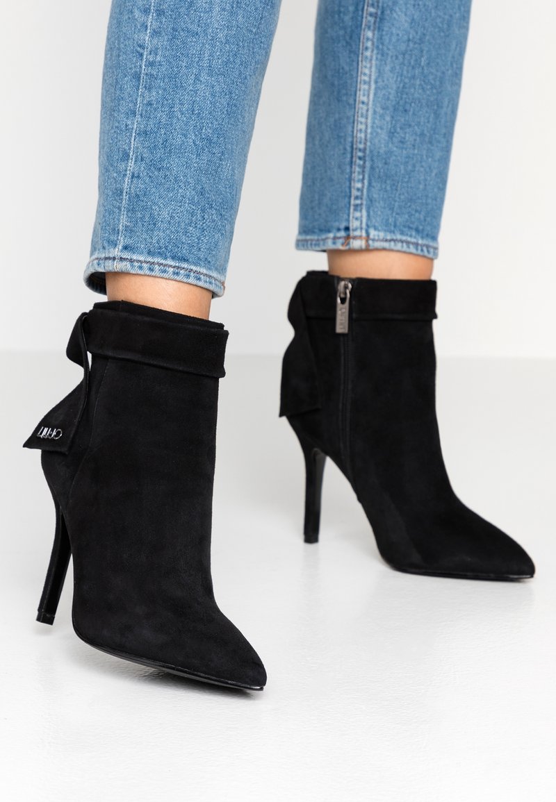 Jambes en jean bleu portant des bottines à talons hauts en daim noir à bout pointu, avec des revers pliés et des fermetures éclair latérales.