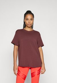 Donna con capelli intrecciati, che indossa una T-shirt larga color bordeaux scuro e leggings Nike Pro rossi vivaci, in piedi contro uno sfondo chiaro e neutro.