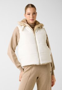Witte gewatteerde bodywarmer met een capuchon afgezet met bont, met een ritssluiting aan de voorkant en een zachte textuur. Draagt over een beige top en bijpassende broek.