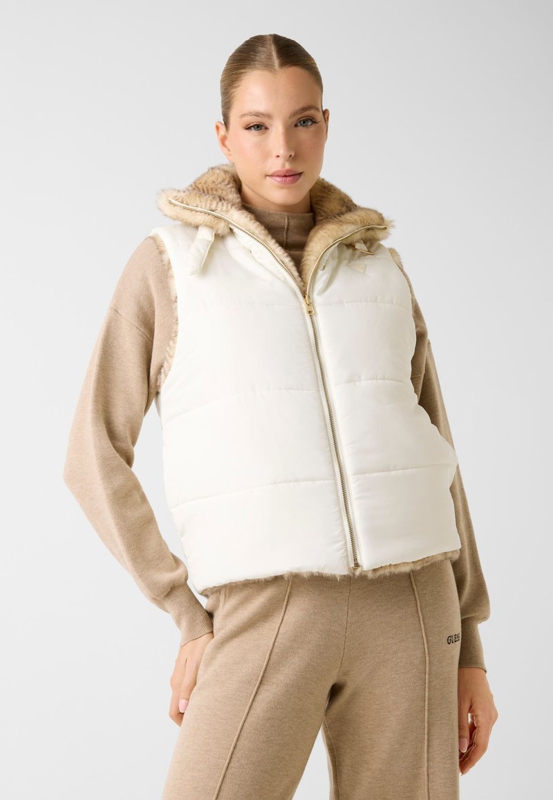 Witte gewatteerde bodywarmer met een capuchon afgezet met bont, met een ritssluiting aan de voorkant en een zachte textuur. Draagt over een beige top en bijpassende broek.