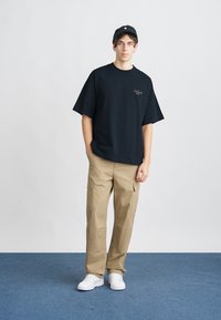 T-shirt nera a maniche corte con un piccolo logo, abbinata a pantaloni cargo color khaki con più tasche e sneakers bianche.