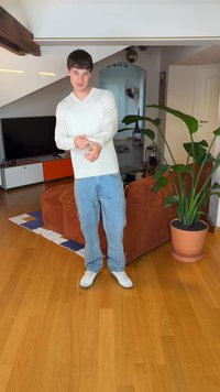 Model v bielej košeli s dlhým rukávom a svetlomodrých rifliach stojí na drevenej podlahe vedľa hnedej manšestrovej pohovky a zelenej rastliny.