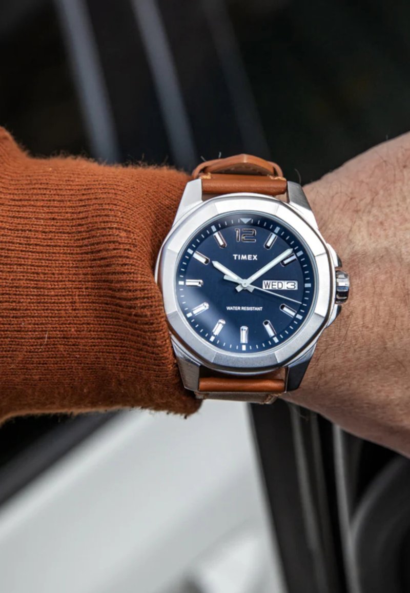 Timex-Uhr mit silbernem Gehäuse, blauem Zifferblatt und braunem Lederarmband. Mit klaren Stundenmarkierungen, Datumsanzeige und Wasserdichtigkeit.