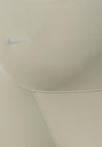 Leggings de sport beige clair en matériau extensible, arborant un logo Nike bleu discret et des coutures flatlock pour le confort.
