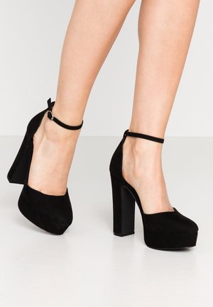 Schwarze Wildleder-Pumps mit runder Spitze, offenen Seiten und einem Knöchelriemen. Sie besitzen eine auffällige Plattform und ein Blockabsatz-Design.