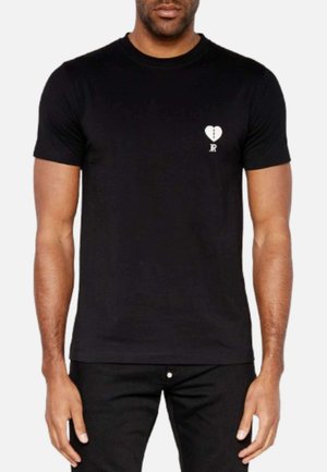 T-shirt nera in cotone a maniche corte con una piccola grafica di un cuore spezzato bianco e dettaglio "JR" sul lato sinistro del petto. Vestibilità classica, texture liscia.