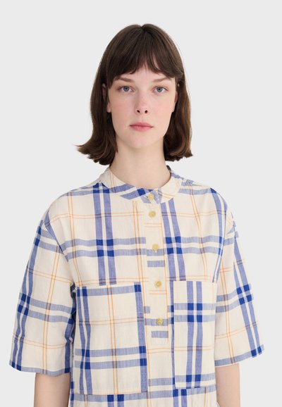Femme aux cheveux courts bruns portant une chemise ample à manches courtes avec un motif à carreaux bleu et orange.