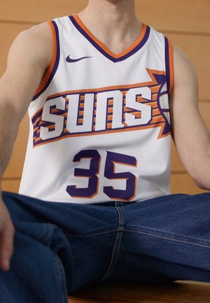 Person iført en hvid Phoenix Suns basketballtrøje nummer 35 med lilla og orange accenter, siddende med benene over kors iført blåt cowboybukser.