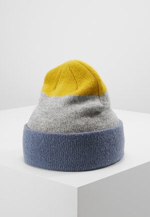 Bonnet tricoté jaune, gris et bleu avec un revers, présenté sur un socle blanc devant un fond uni gris clair.