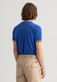 Niebieska koszulka polo z krótkim rękawem, białym kołnierzykiem i prążkowanymi mankietami; w zestawie z beżowymi spodniami z tylnymi kieszeniami.