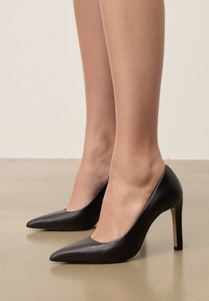 Tacones altos negros de punta afilada, usados sin calcetines, sobre un suelo liso de color claro y con un fondo neutro.