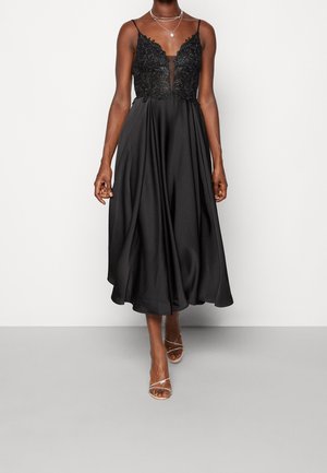 Robe midi noire avec corsage en dentelle et fines bretelles, portée avec des colliers argentés superposés et des talons ouverts à brides.
