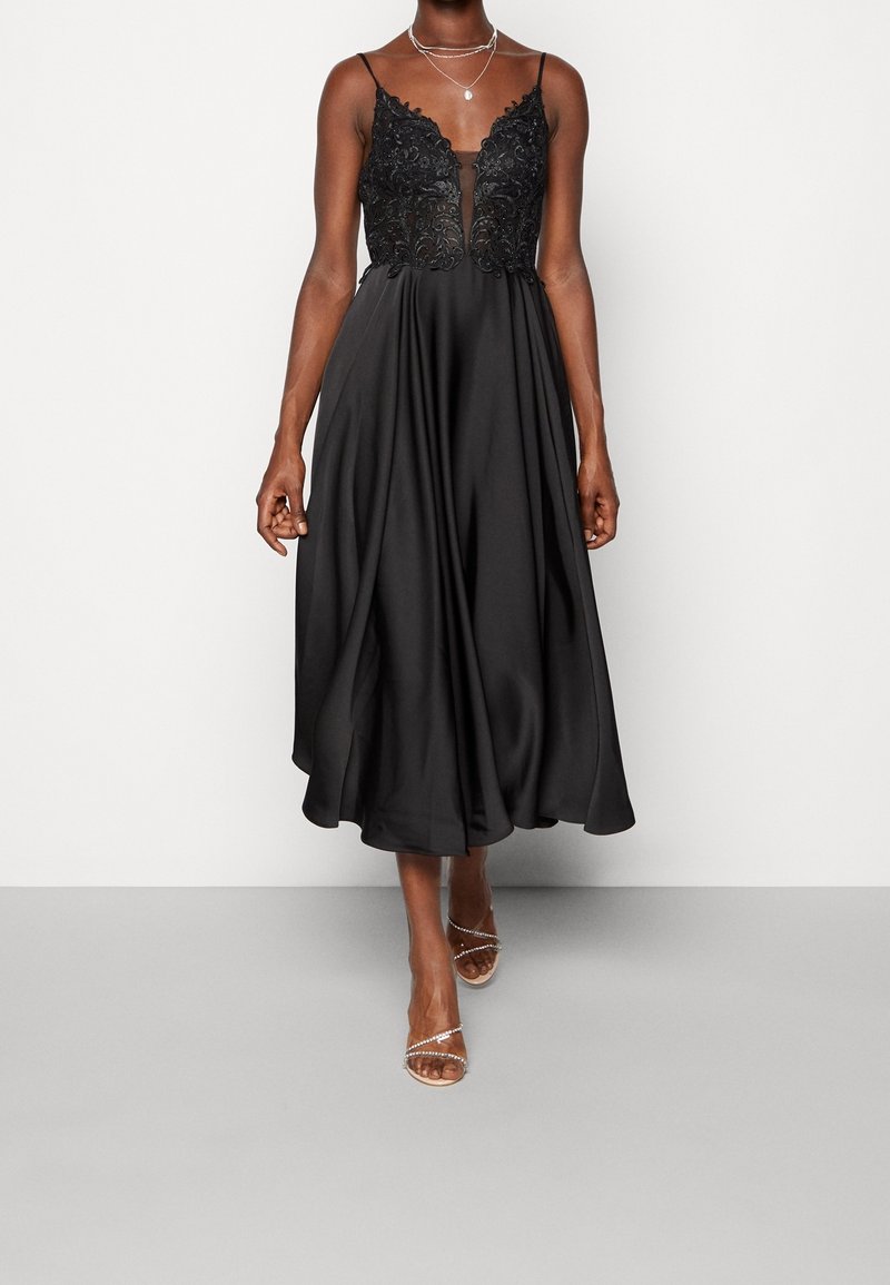 Robe midi noire avec corsage en dentelle et fines bretelles, portée avec des colliers argentés superposés et des talons ouverts à brides.