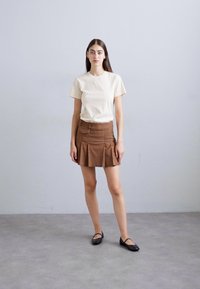 Young woman standing indoors wearing a beige short-sleeve shirt, brown pleated mini skirt, and black Mary Jane flats.