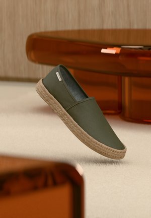 Πράσινο ελαιολάδι παπούτσι τύπου slip-on με πλεκτή σόλα από γιούτα να αιωρείται πάνω από ελαφρύ χαλί σε ένα μοντέρνο δωμάτιο με κεχριμπαρένια έπιπλα.