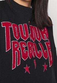 Sudadera negra con texto en purpurina roja brillante "TOUJOURS REBELLE" con contornos blancos, acentuada con estrellas y rayos rojos.