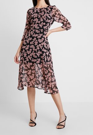 Mujer con vestido midi negro con estampados florales rosas y mangas tres cuartos transparentes, combinado con tacones altos negros con tiras.