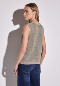 Ljust beige stickad linne utan ärmar med ett texturerat mönster, porerat med mörkblå denimjeans, sedd bakifrån.