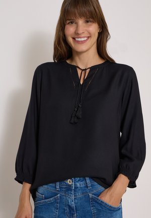 Femme souriante portant un chemisier noir à manches longues avec des liens à pompons et un jean bleu, main dans la poche.