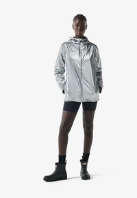 Mujer con chaqueta de lluvia plateada con capucha, pantalones cortos de ciclismo negros, calcetines negros y botas de lluvia negras hasta el tobillo, de pie con las manos en los bolsillos.